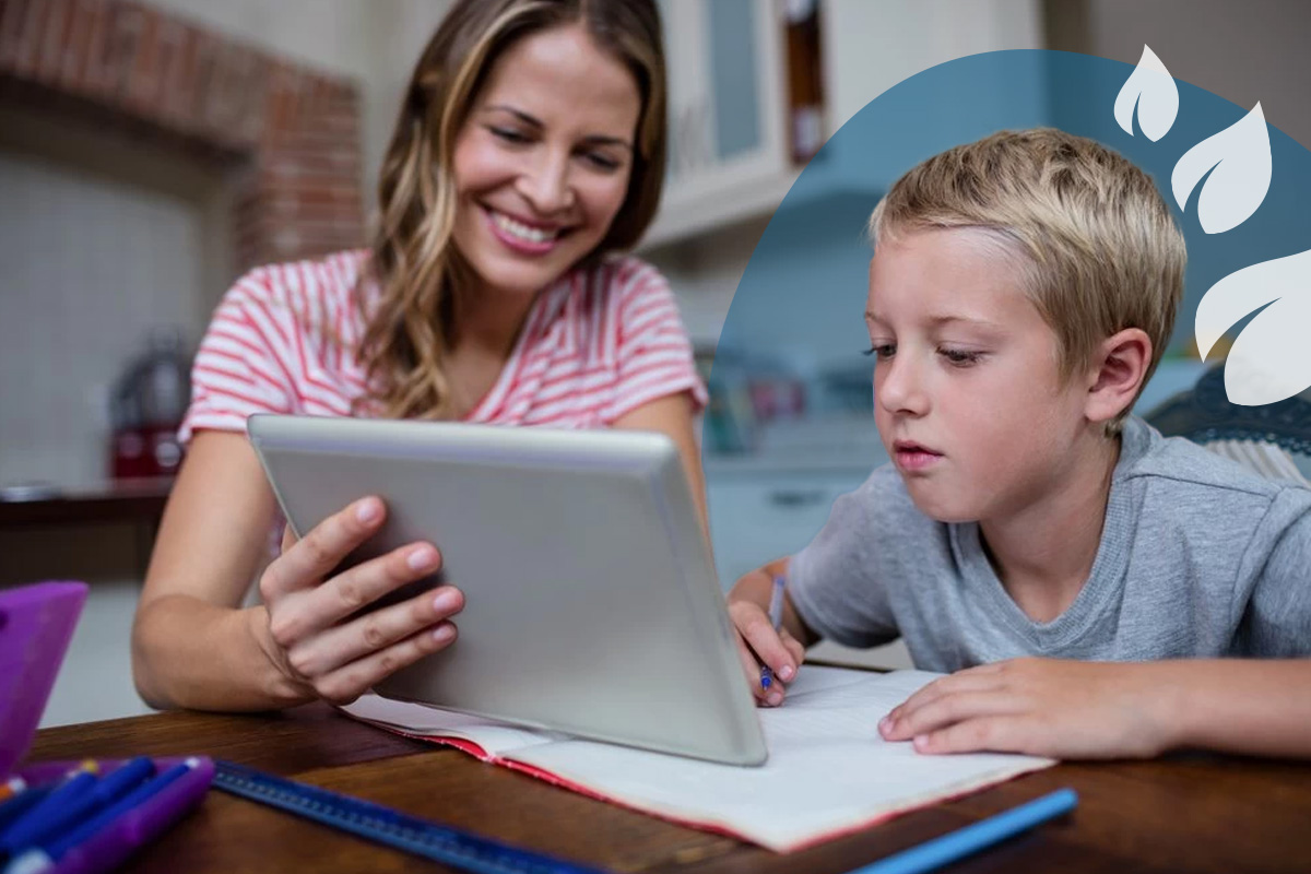 A homeschooling mum and son sitting at a desk. The mum is smiling and showing her son something on a tablet device, which the boy is studying intently.