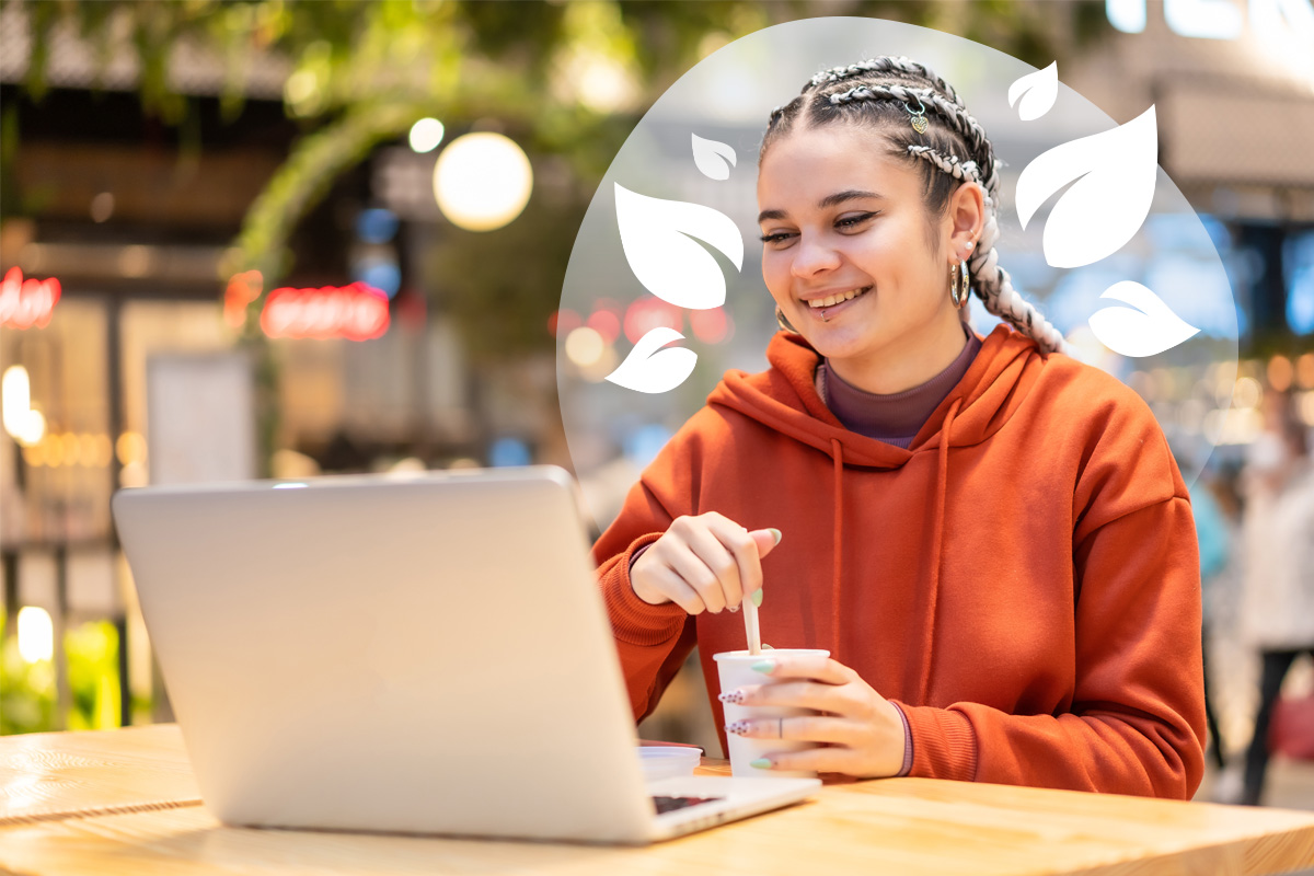 A young person with braided hair smiling while working on a laptop and holding a drink cup in a bright indoor setting.