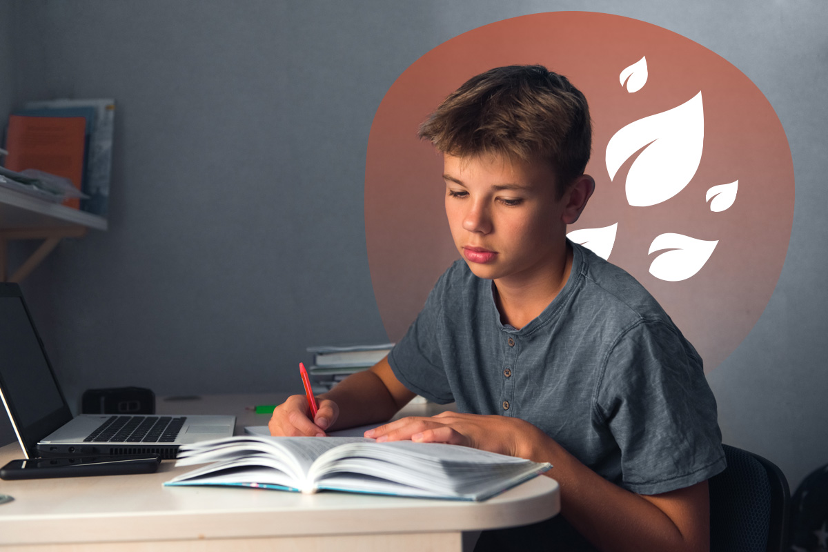 Teen boy studying at a desk, writing in an open book beside a laptop.