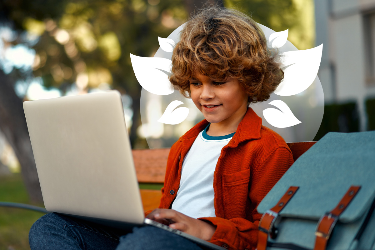 A young student with curly hair, sitting on a bench outdoors and smiling while using a laptop.