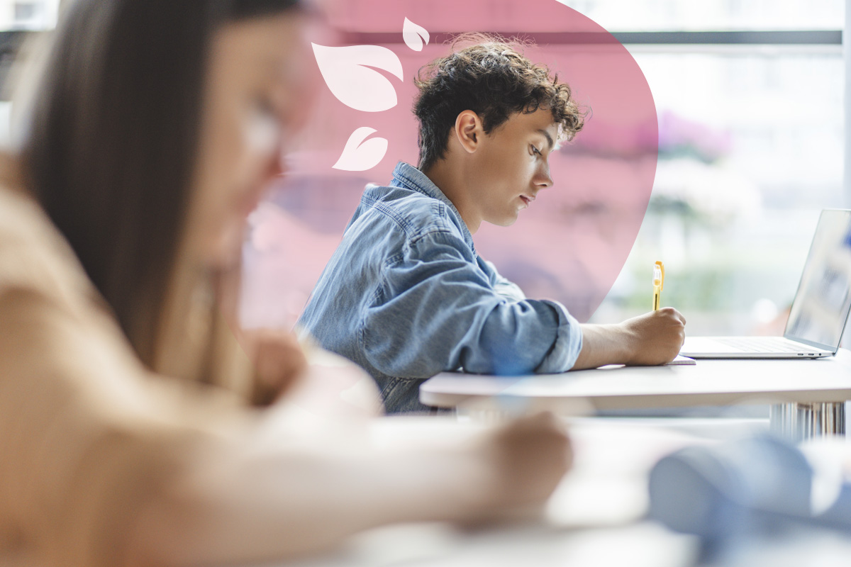 A young student with curly hair is focused on writing during an exam or study session, with a laptop nearby.
