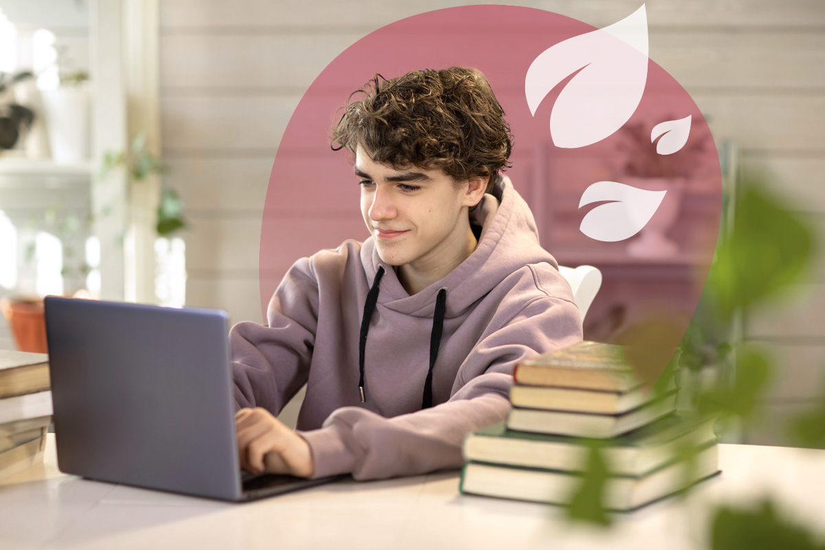 A young student with curly hair, wearing a hoodie, smiling slightly while working on a laptop at a desk with stacked books.