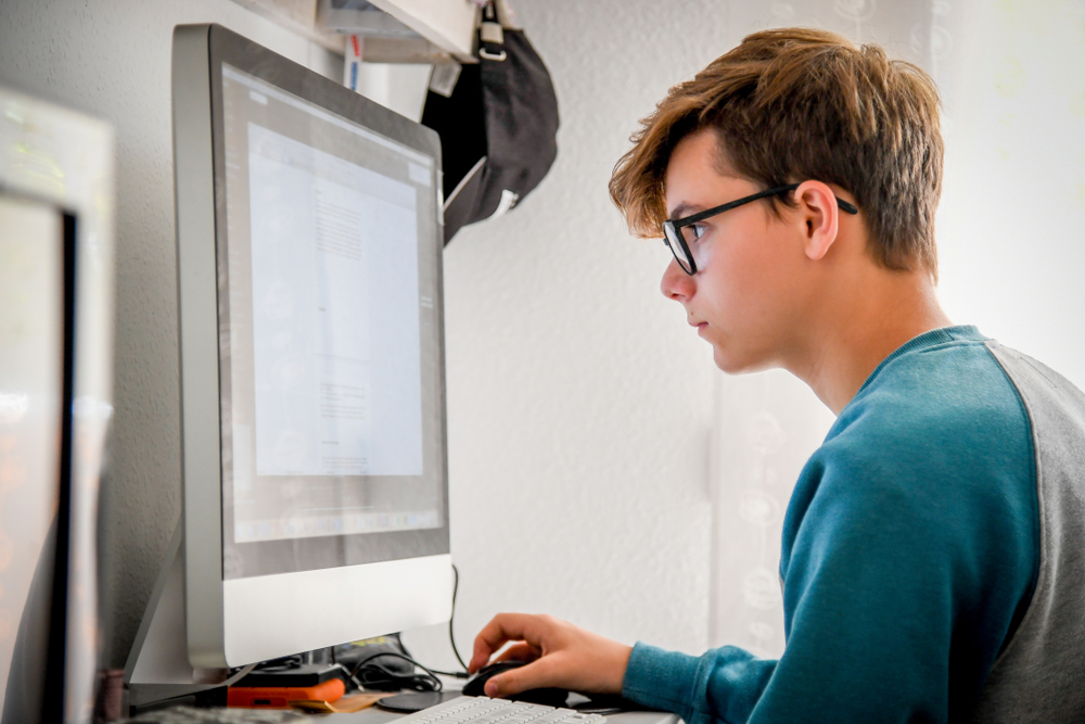 Focused teenage boy using a desktop computer for study or research in a home learning space.