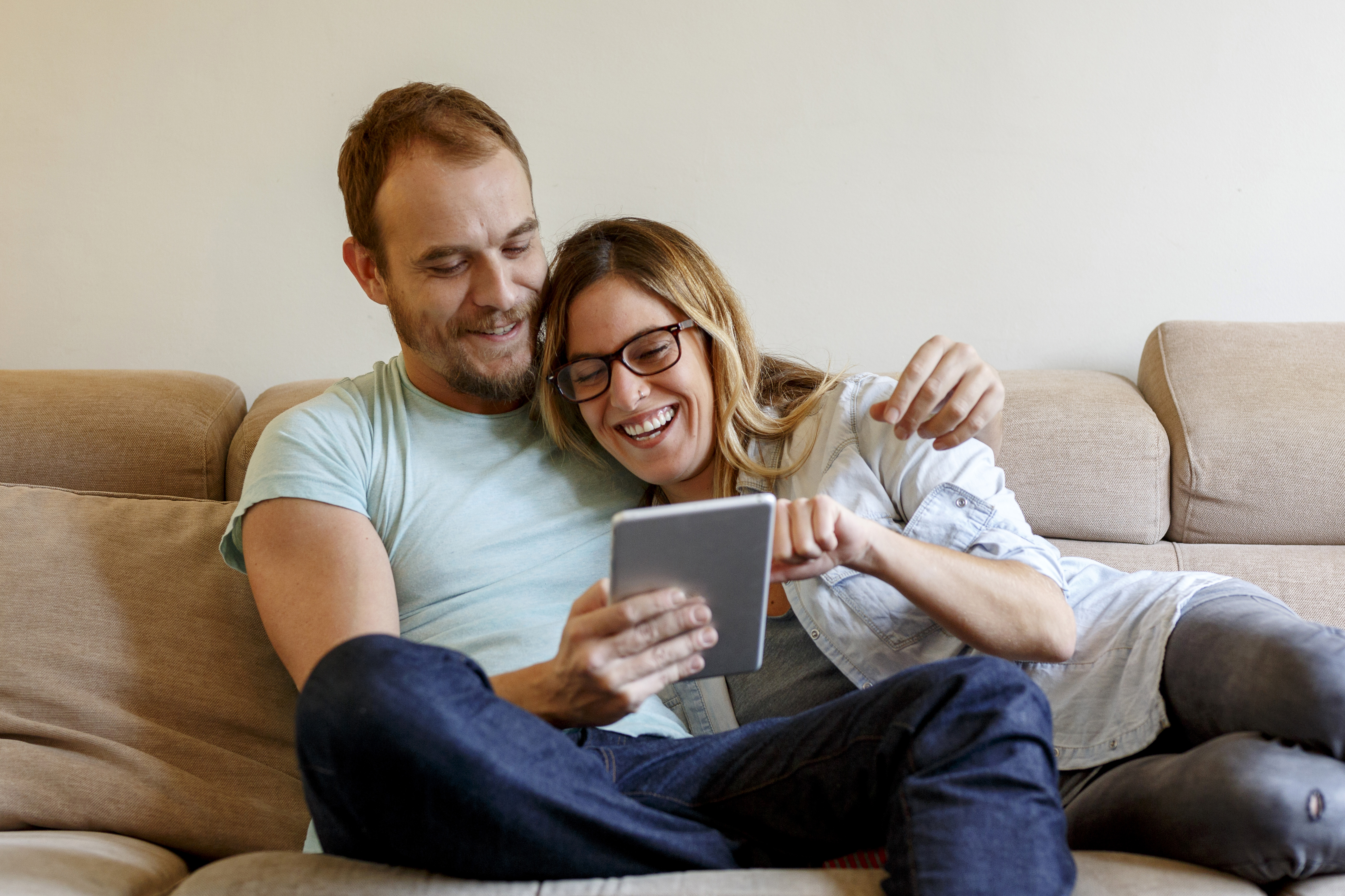 Happy couple sitting on sofa, laughing together while viewing a tablet.