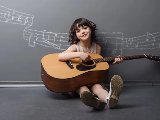Smiling girl seated on the floor playing an acoustic guitar with chalk music notes drawn on the wall behind.
