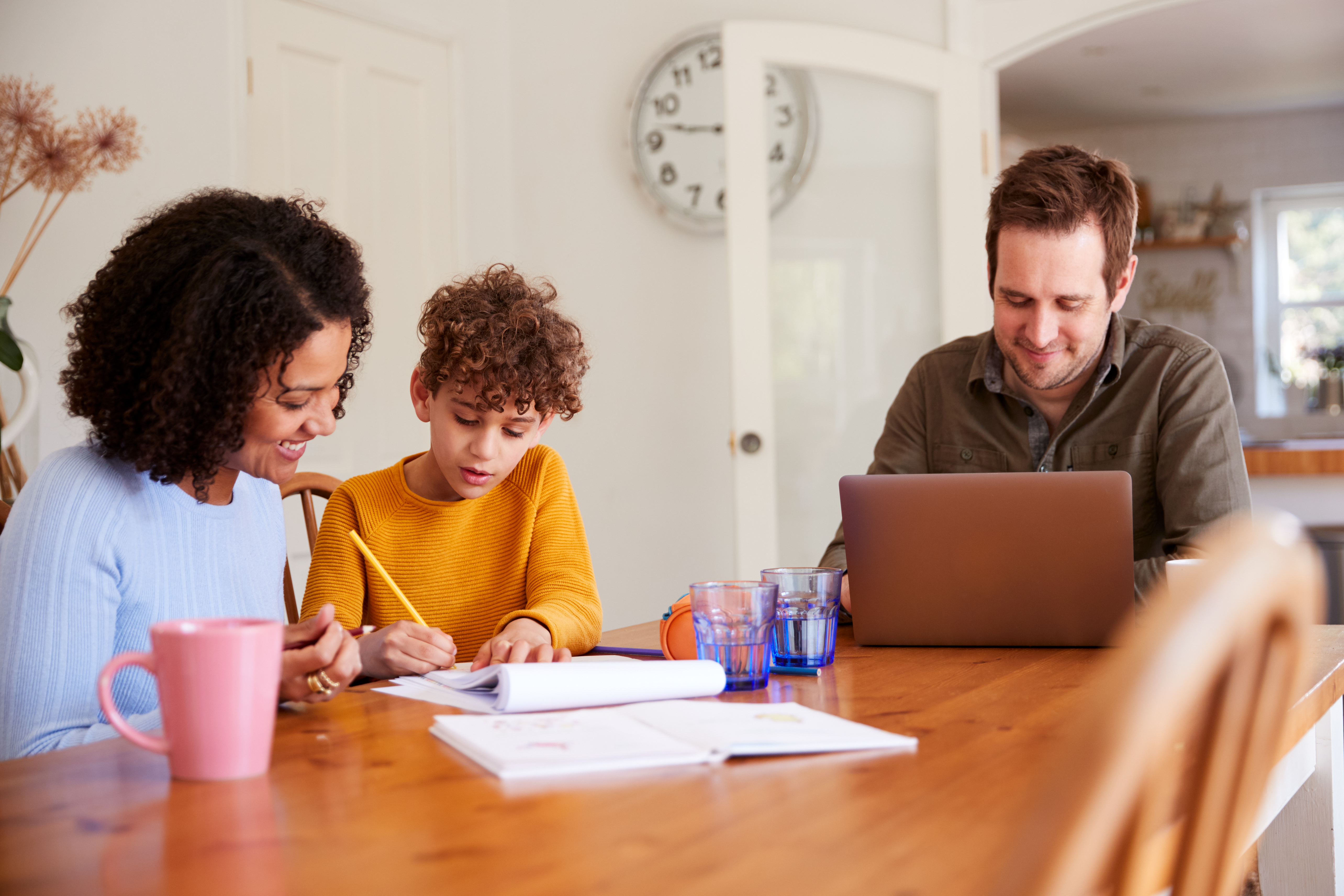 Mother helping child with work at a table while father works on a laptop nearby