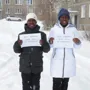 Two individuals holding signs with their names, standing in a snowy environment in front of buildings.