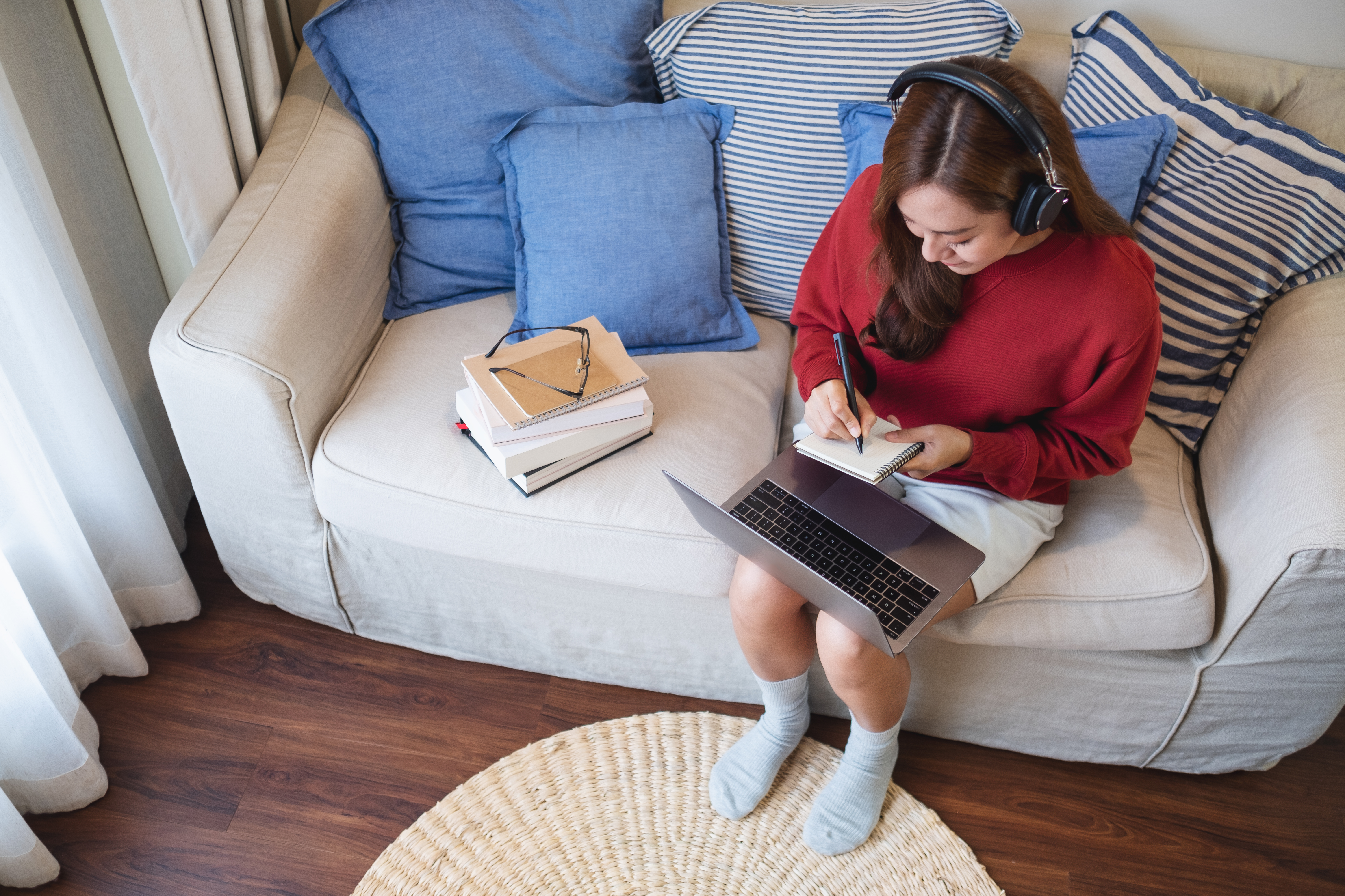 Teenage girl writing notes with laptop on lap and headphones on, sitting on a sofa with study materials beside her