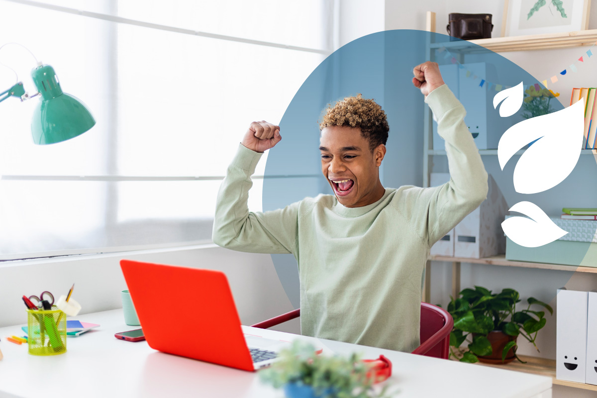 Teen boy celebrating success in front of a red laptop at a tidy home study desk.