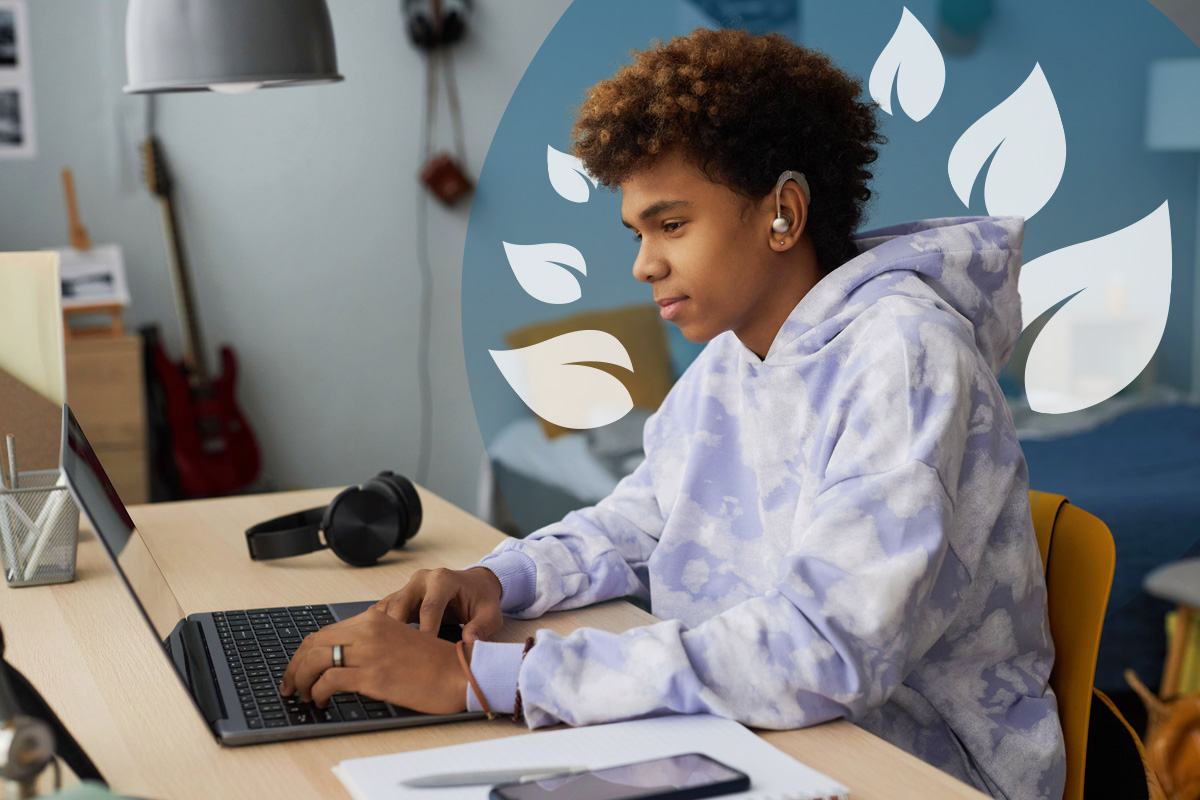 Focused boy wearing a hearing aid, studying at his desk with a laptop and notebook.