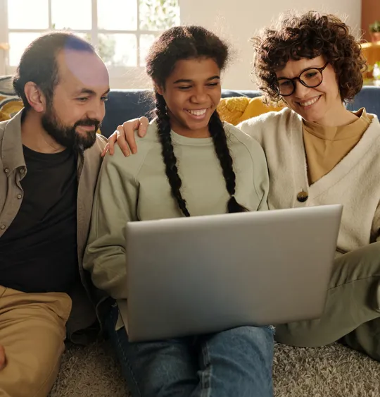 A smiling mother, father, and their teenage daughter sit together on the living room floor, happily watching something on a laptop.