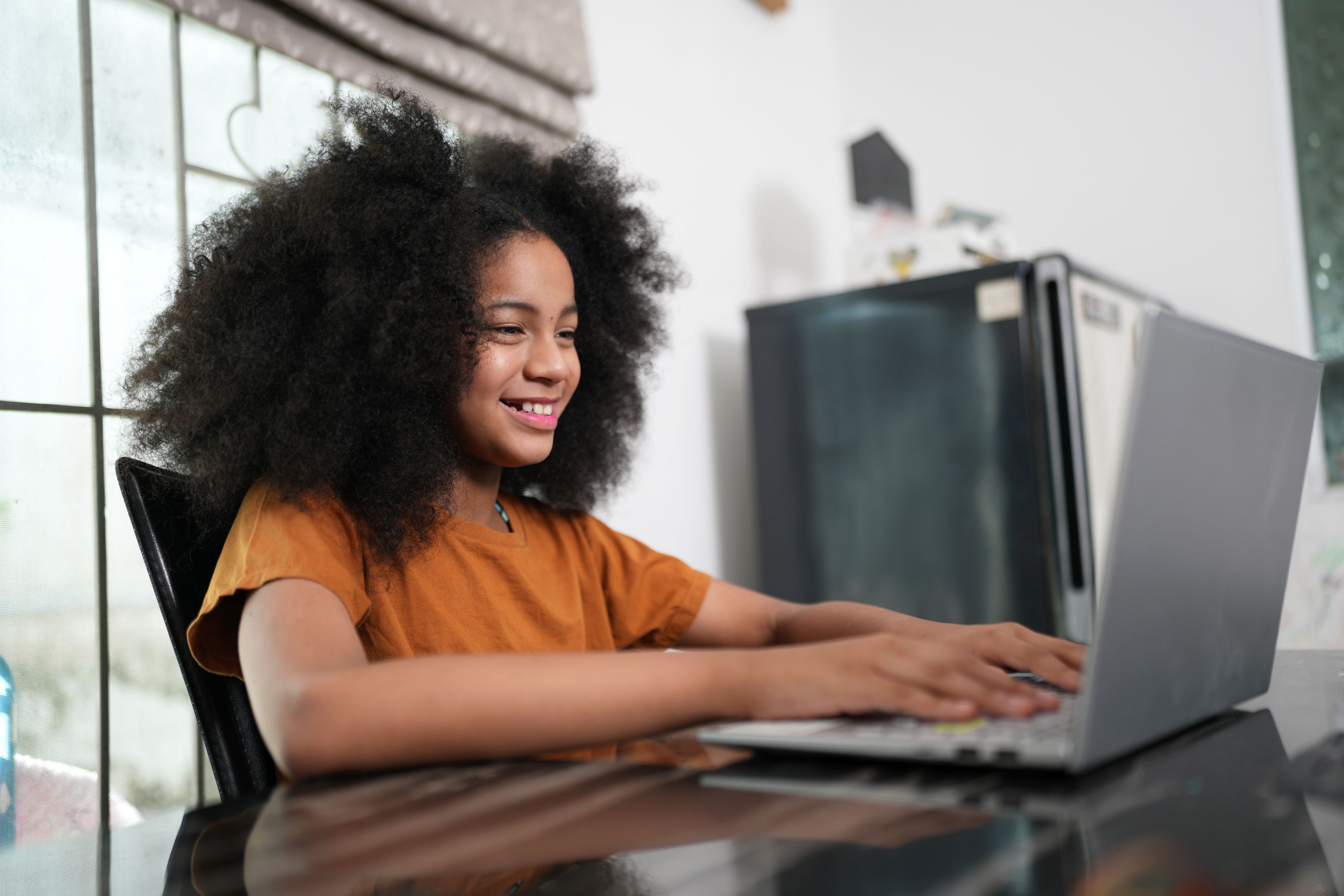 Young girl smiling as she types on a laptop at a kitchen table.