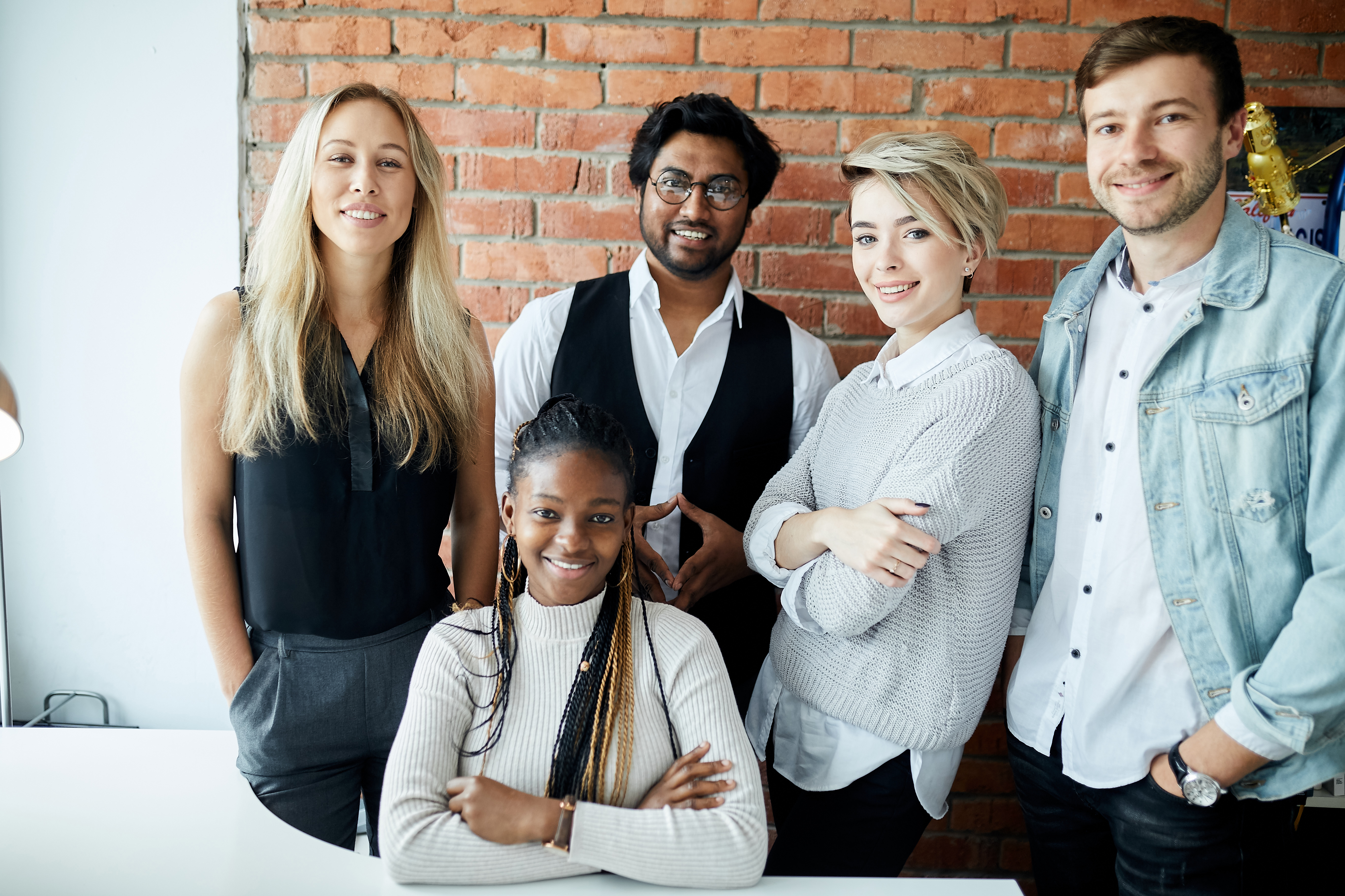 Group of five young professionals smiling together in a modern office with brick walls.