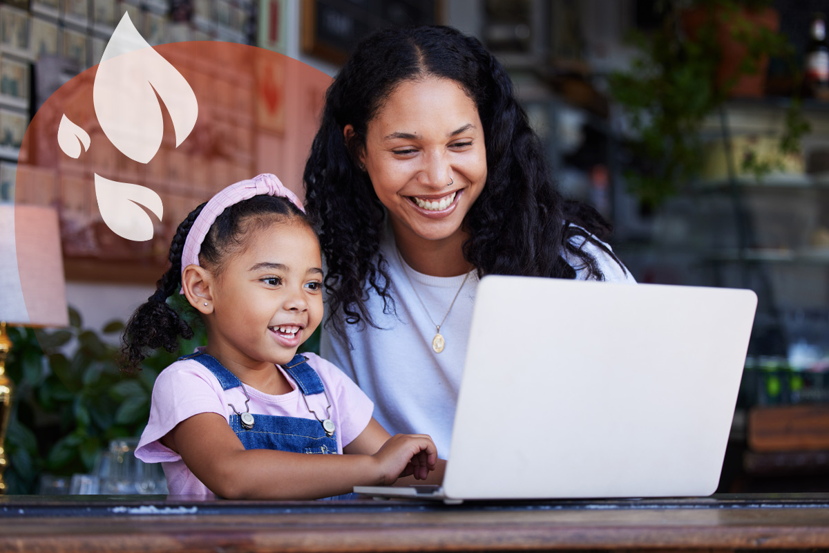 An adult and a young girl smiling and looking at a laptop together, appearing to be in a cafe setting.