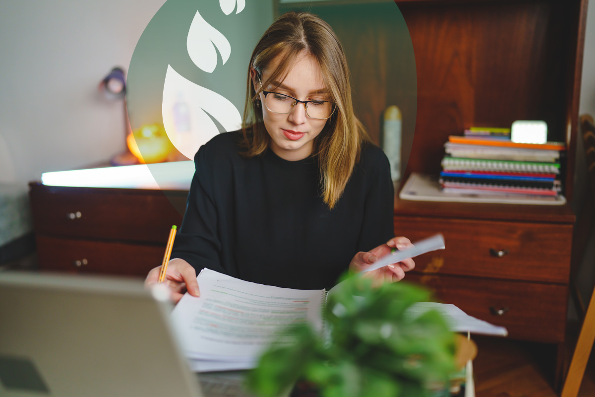 A young woman with glasses holding papers and writing, seated at a desk with a laptop and a plant, with a green leafy design in the background.