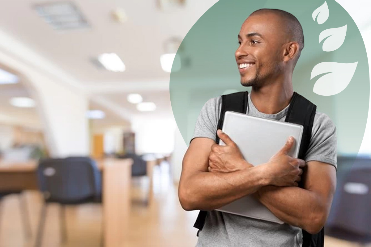 A young man with a black rucksack smiling to the left, holding a laptop to his chest with both arms.
