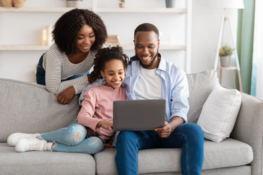 A homeschooling mum, dad and young daughter in their living room smiling at a laptop. The dad and daughter are sat on the sofa, whilst the mother is leaning on the back of it.