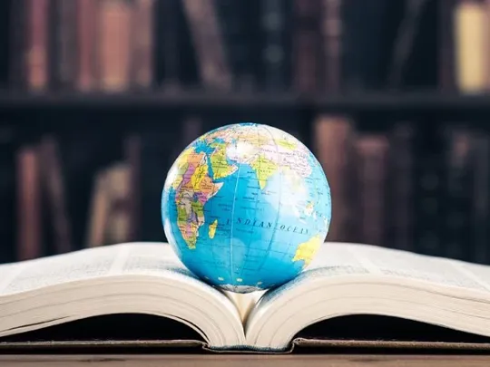 A large book within a library open at the middle with a globe laid on top of it.