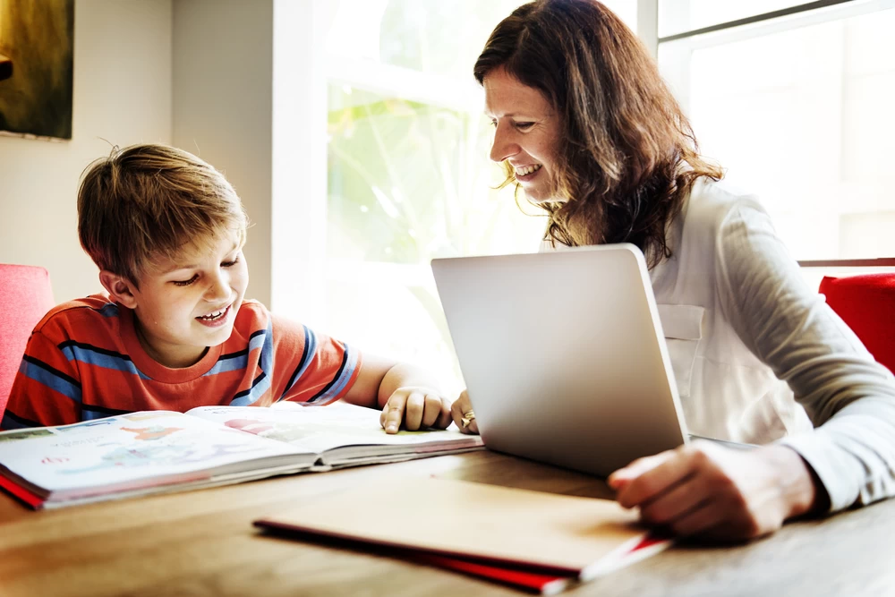 A mum helping her young son homeschool. They are both sat smiling at a desk with a laptop and a textbook.
