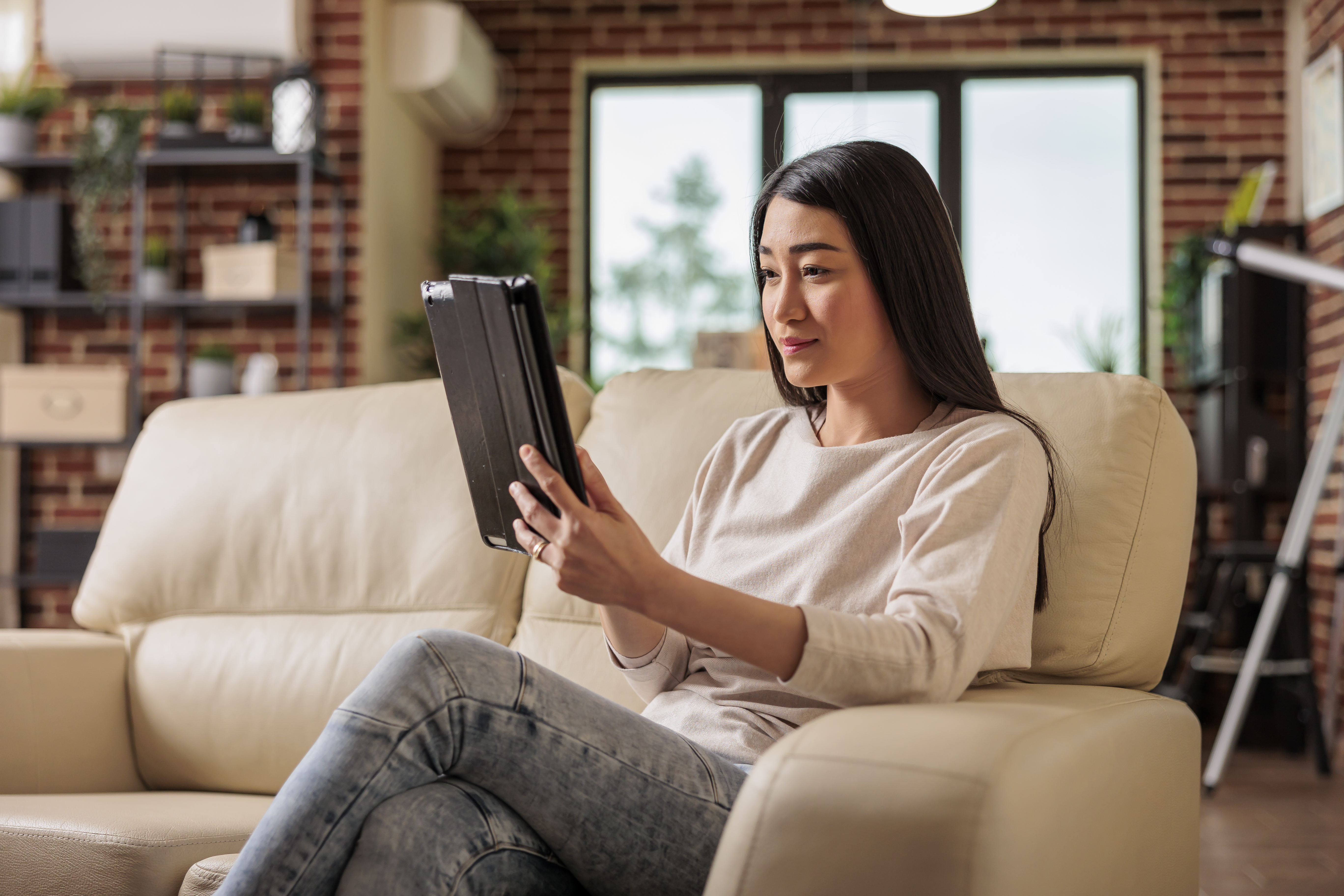 Young woman on cream sofa focused on a tablet in a modern living room setting.
