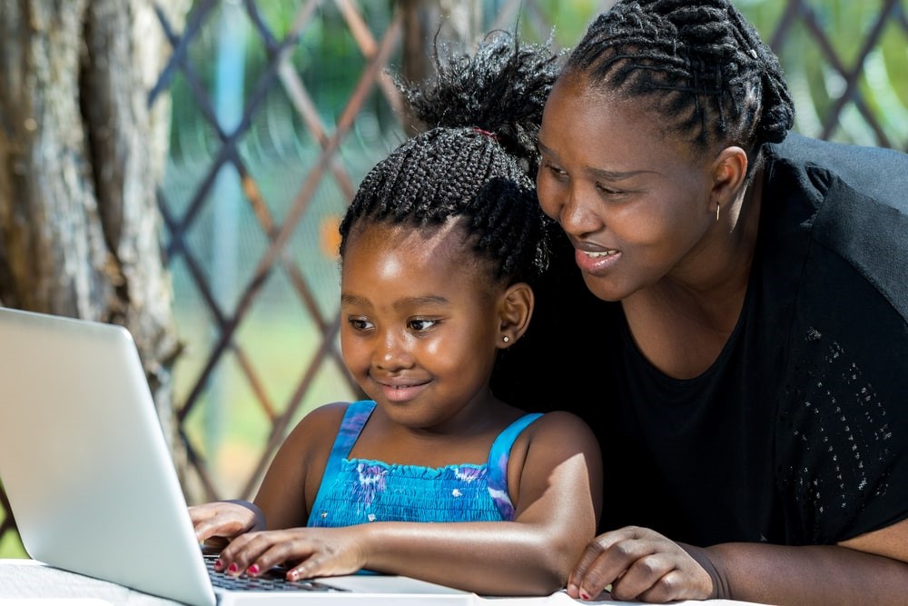 dult assisting a young girl with her studies on a laptop outdoors, as part of their South Africa home education programme.