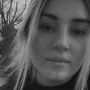 A close up, black and white portrait of a young woman looking directly at the camera, with blurred trees in the background.