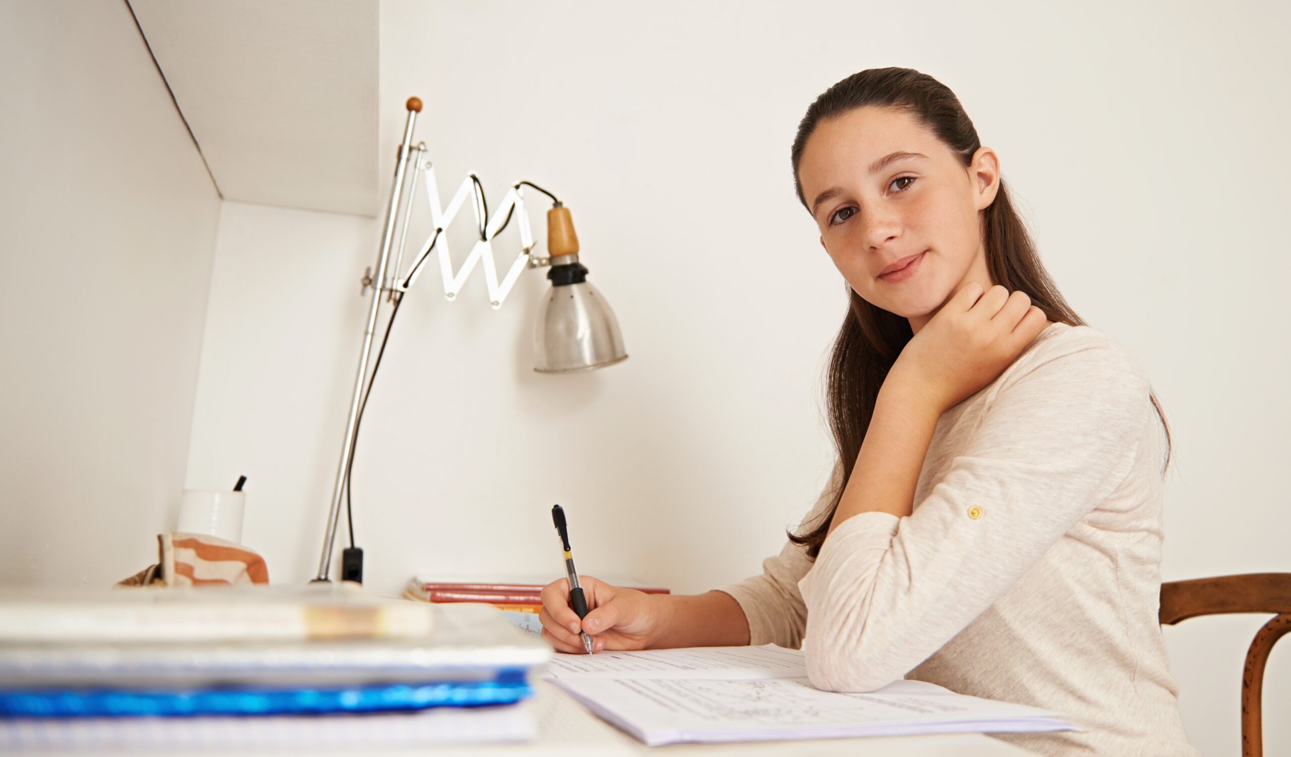Teenage girl writing notes at a desk with a lamp and open books.